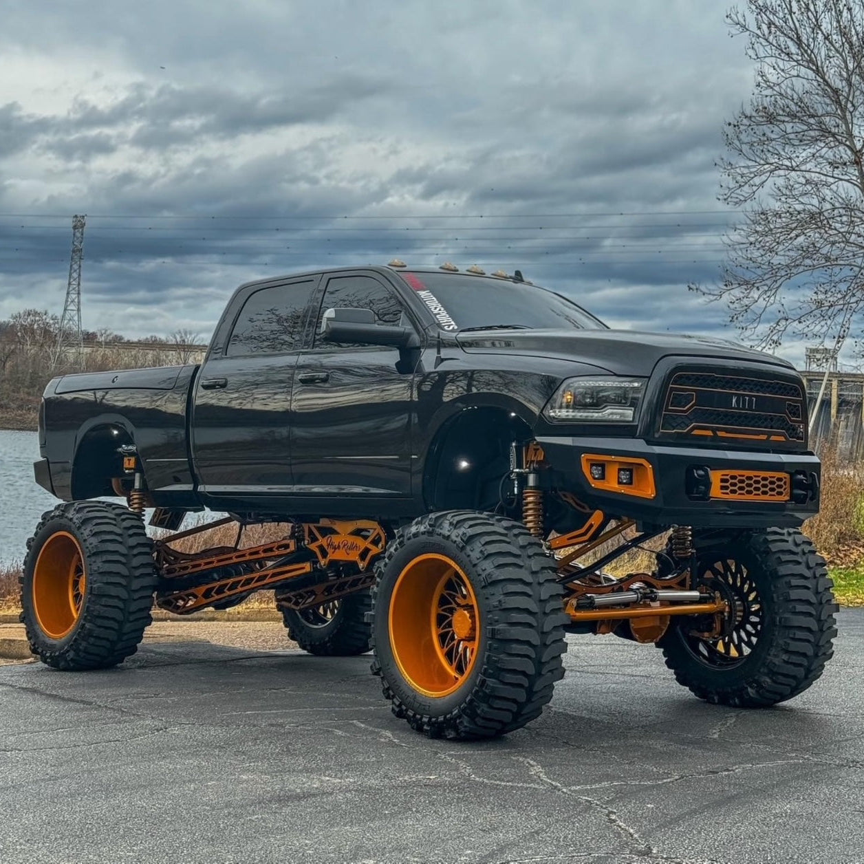 Black pickup truck featuring FLOG front and rear bumpers, large tires, and orange rims on a cloudy day. large tires and orange rims on a cloudy day.